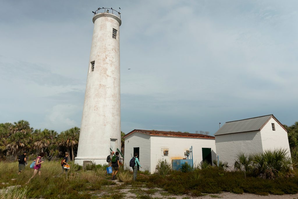 Students in single file line walk past a white lighthouse in an overgrown habitat