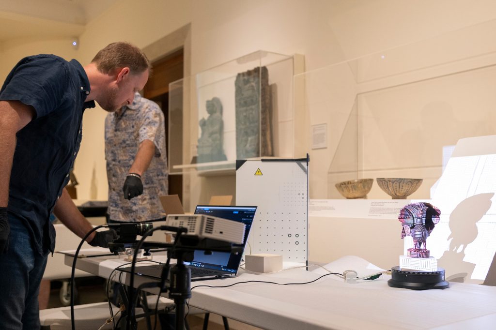 Man wearing gloves looks at computer screen while standing next to a machine and object on display