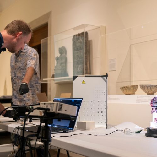 Man wearing gloves looks at computer screen while standing next to a machine and object on display