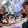 Professor in glasses leans over table to critique a student's work as comics appear on screen behind him