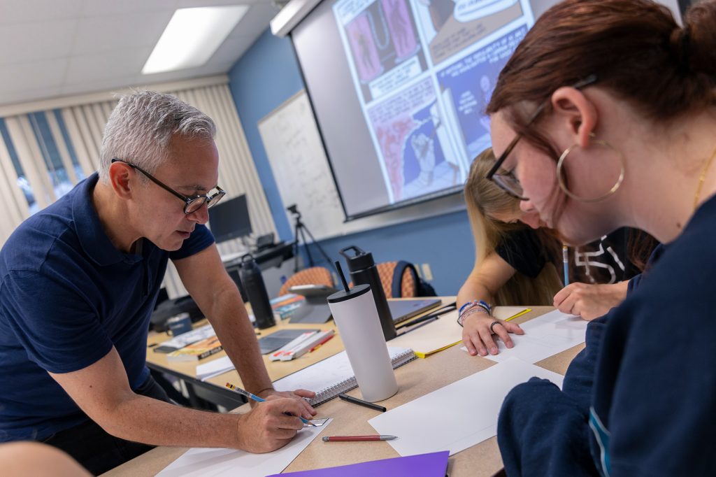 Professor in glasses leans over table to critique a student's work as comics appear on screen behind him