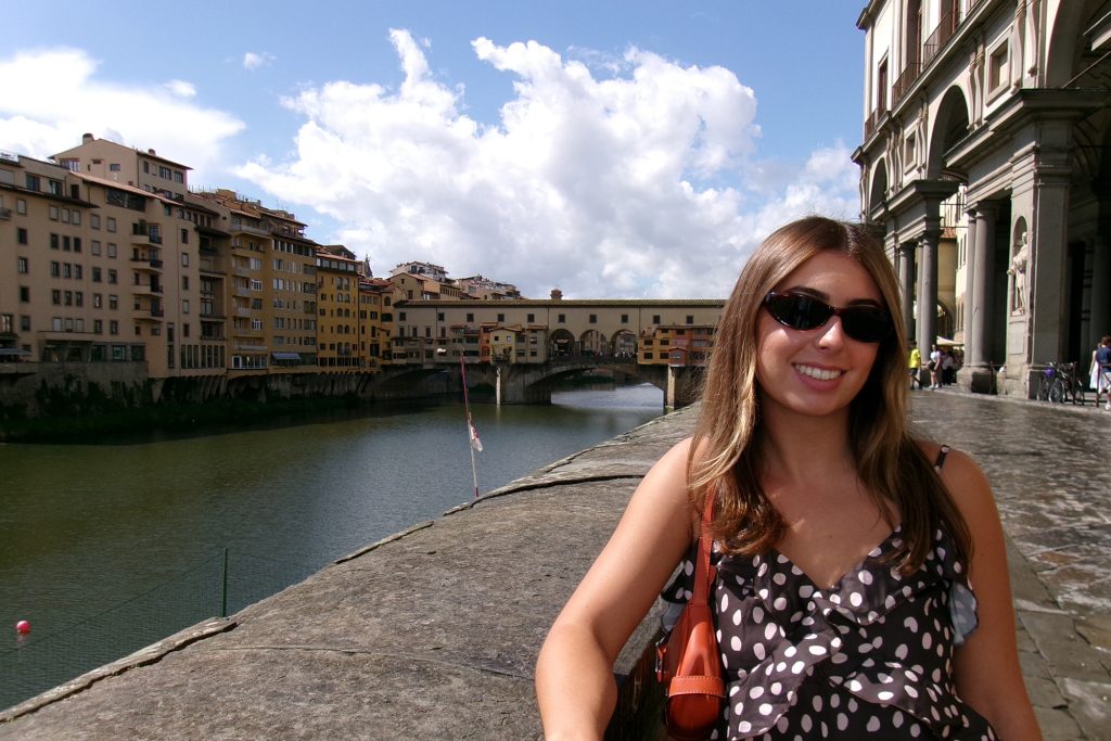 Student wearing purse and sunglasses stands in front of river passing through an old European city