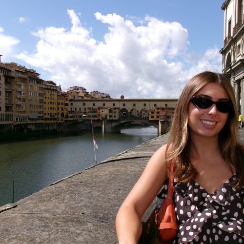 Student wearing purse and sunglasses stands in front of river passing through an old European city