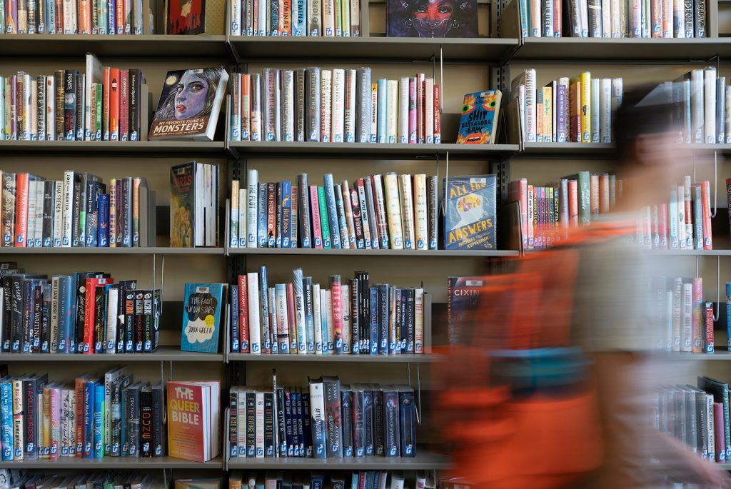 Shelves of books and a blurred image of a student wearing a backpack walking by