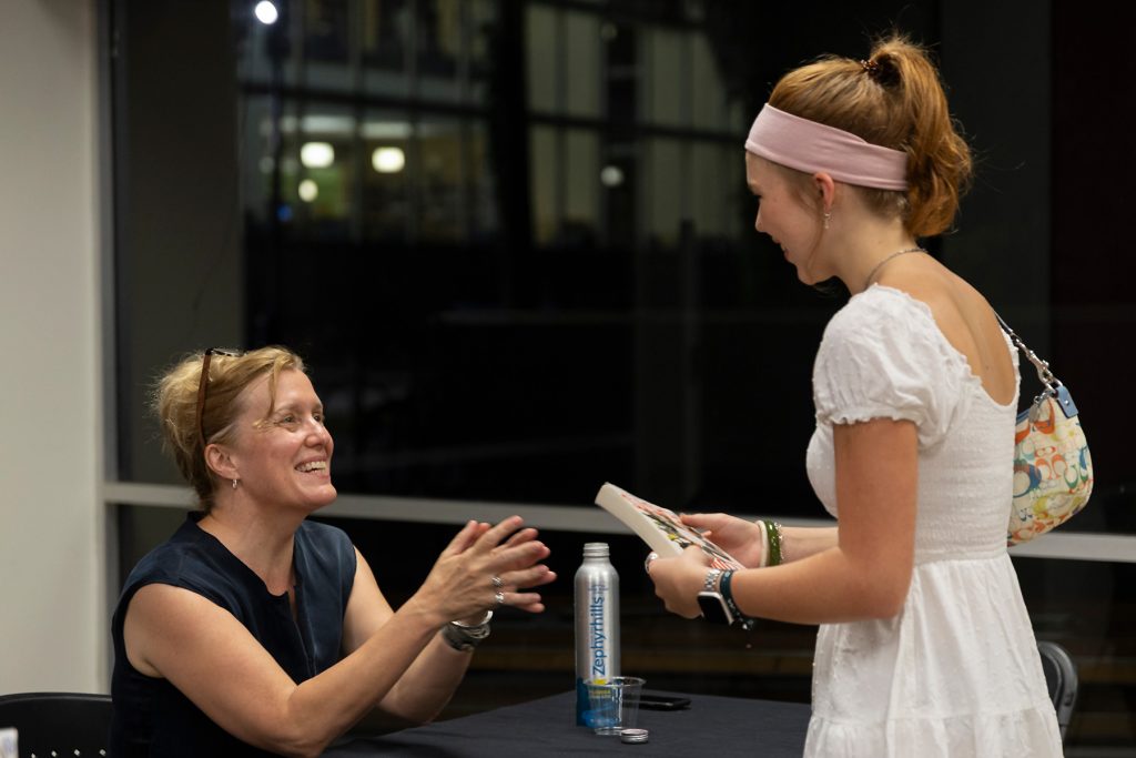 Author seated at table smiling at a student holding book