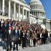 Large group of people including students stand in formal attire in front of the Capitol steps