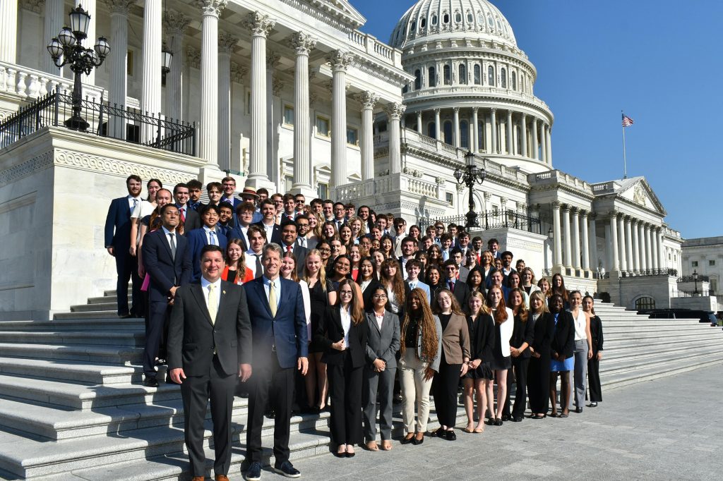 Large group of people including students stand in formal attire in front of the Capitol steps