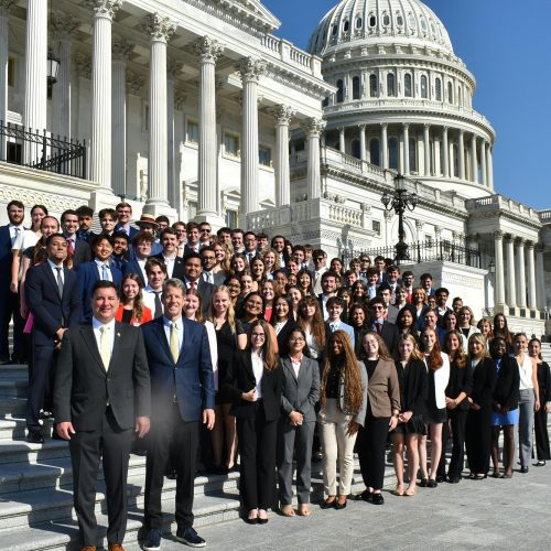 Large group of people including students stand in formal attire in front of the Capitol steps