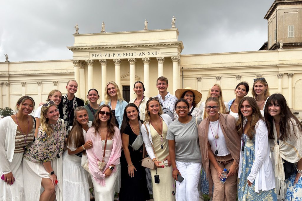 20 people huddled together wearing dresses stand in front of a Vatican museum