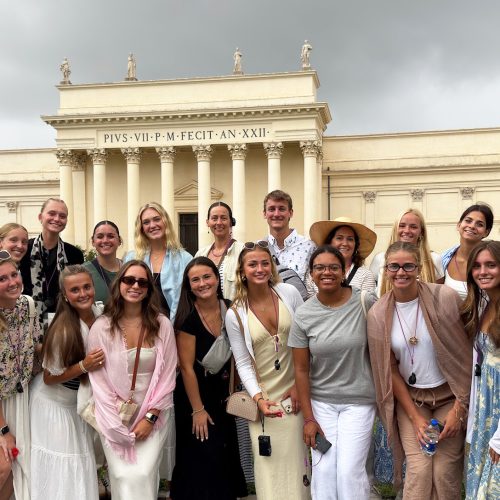 20 people huddled together wearing dresses stand in front of a Vatican museum