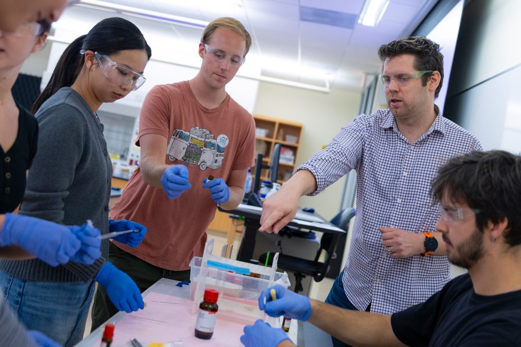 Professor and students in protective eyewear in a lab point to vials on a table