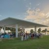 A group of people in formal attire stand under and around an open-air building by the water as the sun sets