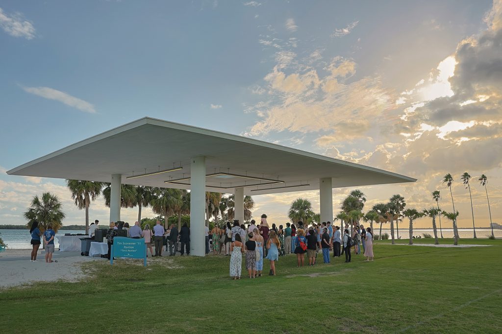 A group of people in formal attire stand under and around an open-air building by the water as the sun sets