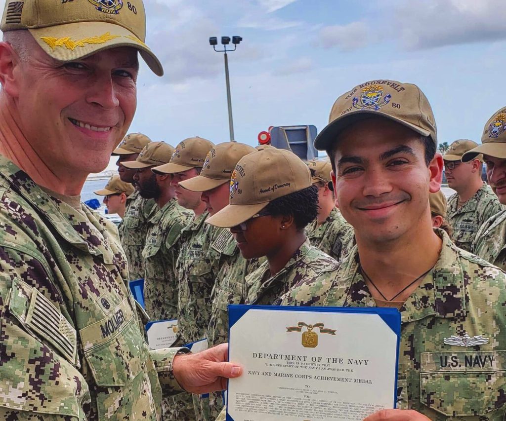 Young man in fatigues holding certificate presented to him by another member of the military
