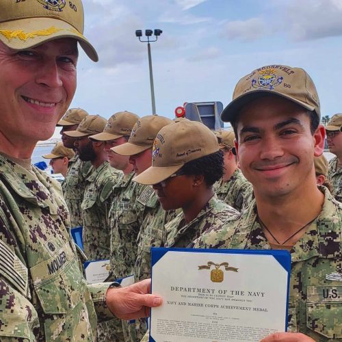 Young man in fatigues holding certificate presented to him by another member of the military