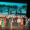 Cast and crew stand on stage in front of the screen showing a scene of a man standing on a dock by a boat