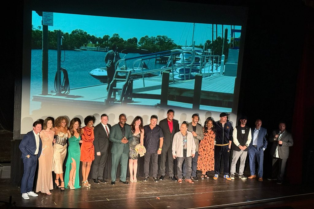 Cast and crew stand on stage in front of the screen showing a scene of a man standing on a dock by a boat