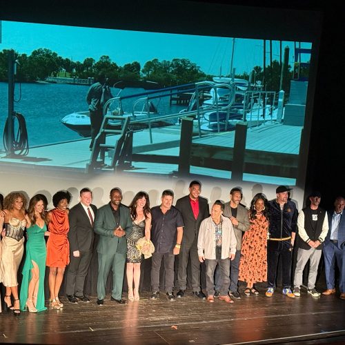 Cast and crew stand on stage in front of the screen showing a scene of a man standing on a dock by a boat