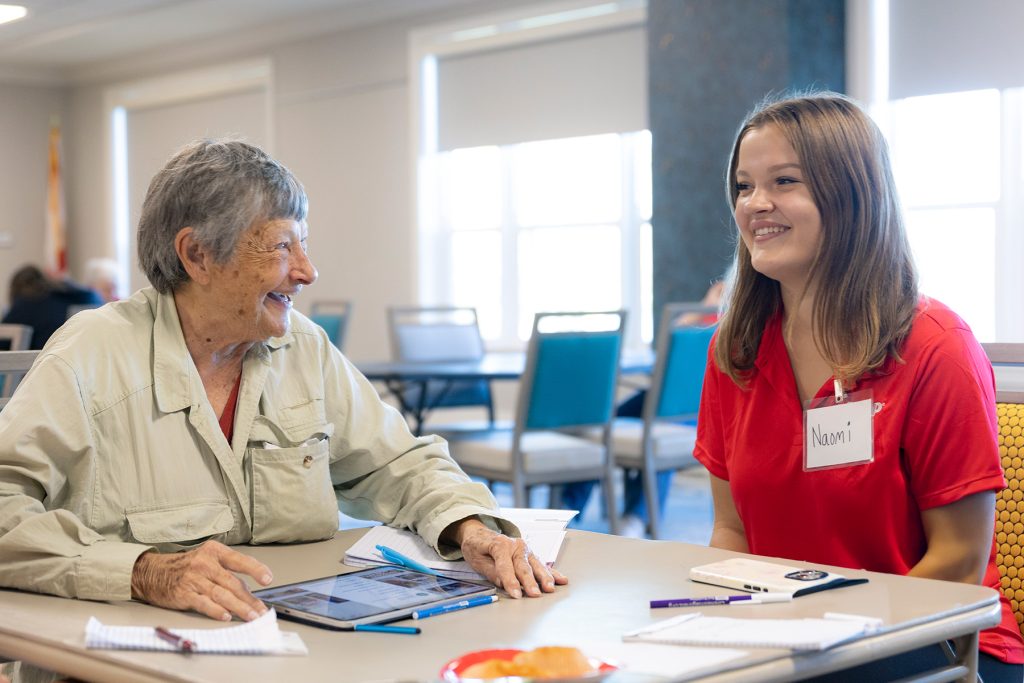 Elderly woman laughs with a college student while seated at a table