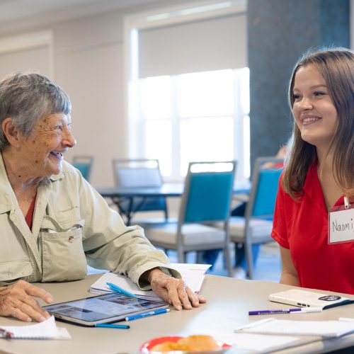 Elderly woman laughs with a college student while seated at a table