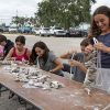 Students sit at a long table covered with oysters shells and string them together on ropes