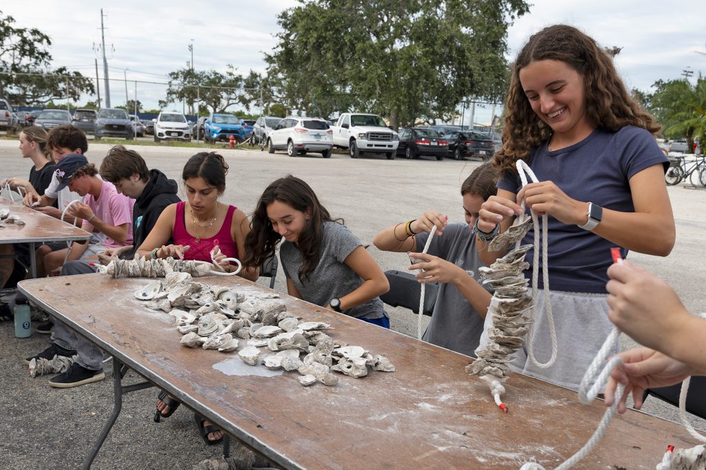 Students sit at a long table covered with oysters shells and string them together on ropes