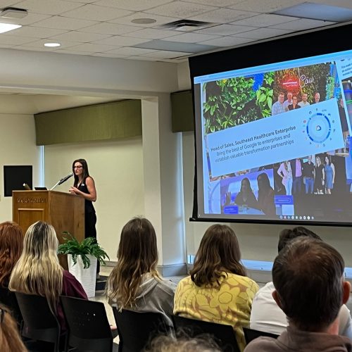 Woman wearing glasses stands at a podium next to a large screen in front of rows of seated students