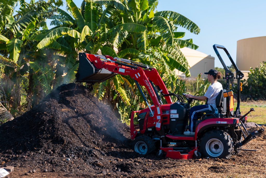 Student sits atop a red tractor as it dumps dirt onto a large pile
