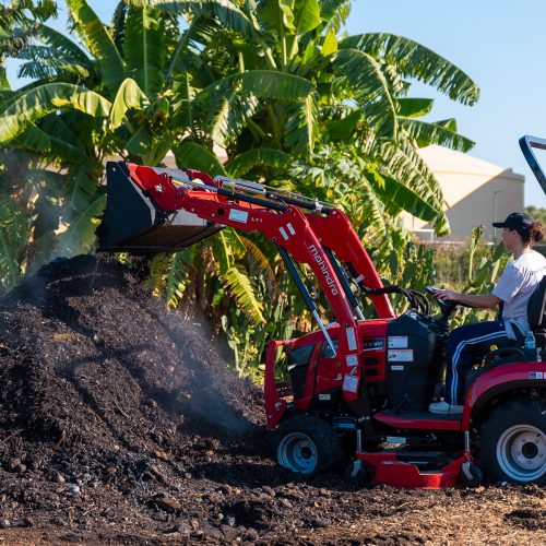 Student sits atop a red tractor as it dumps dirt onto a large pile