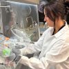 Student in lab jacket and gloves leans over table where she uses scientific equipment