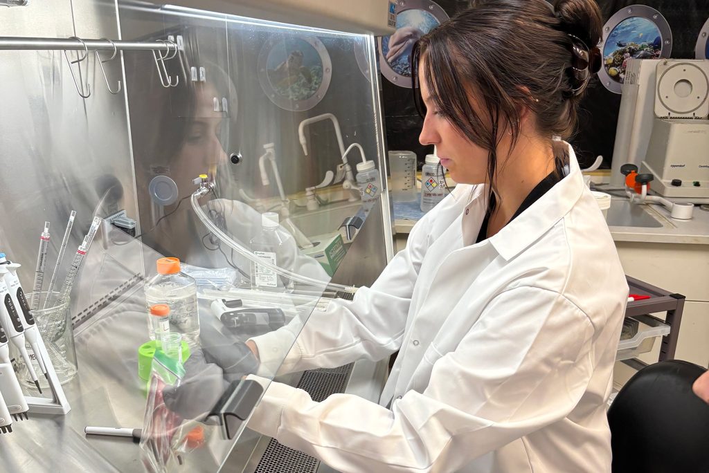 Student in lab jacket and gloves leans over table where she uses scientific equipment