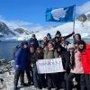 Group of students and professor huddle together while holding a flag and a banner that reads THANK YOU while standing in front of mountains covered in glaciers