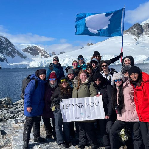 Group of students and professor huddle together while holding a flag and a banner that reads THANK YOU while standing in front of mountains covered in glaciers