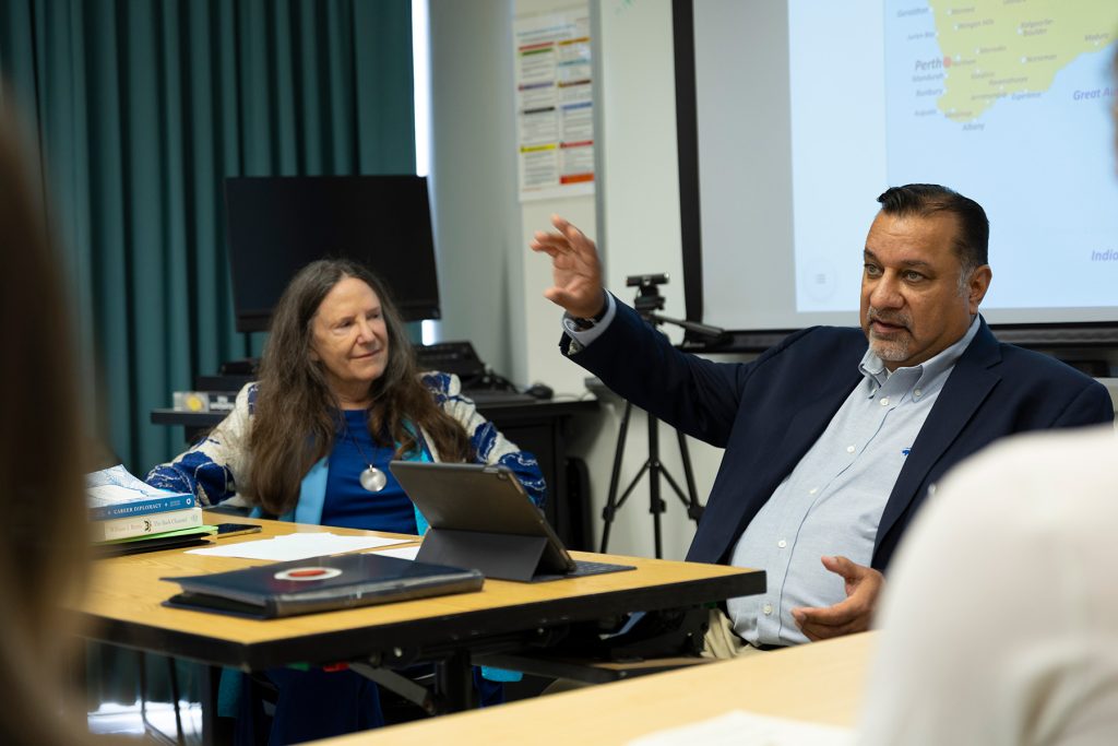Man in dress jacket sits next to professor in front of screen showing a world map and speaks to students