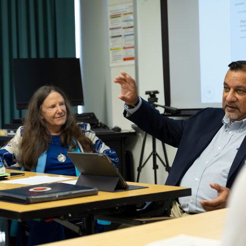Man in dress jacket sits next to professor in front of screen showing a world map and speaks to students
