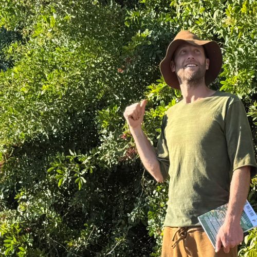 Man in hat with large brim and green and brown clothes stands in front of large bush while pointing to it and holding a notebook