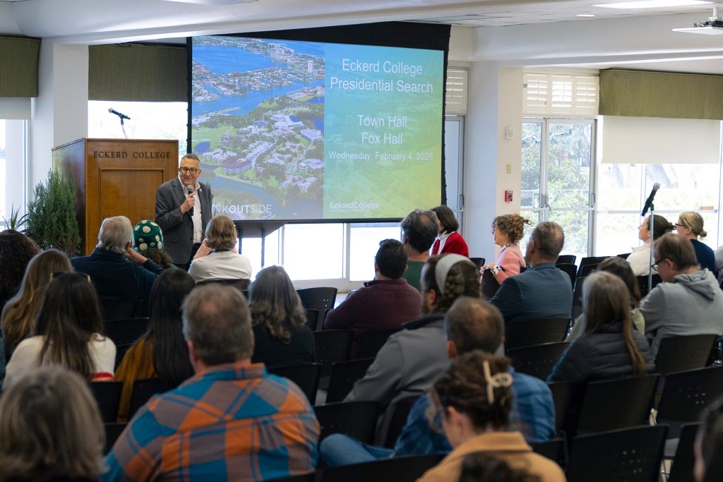 Man in suit stands in front of a large crowd while holding a microphone