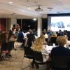Man at podium stands next to large screen as an audience seated at large round tables listens