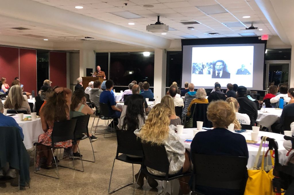 Man at podium stands next to large screen as an audience seated at large round tables listens