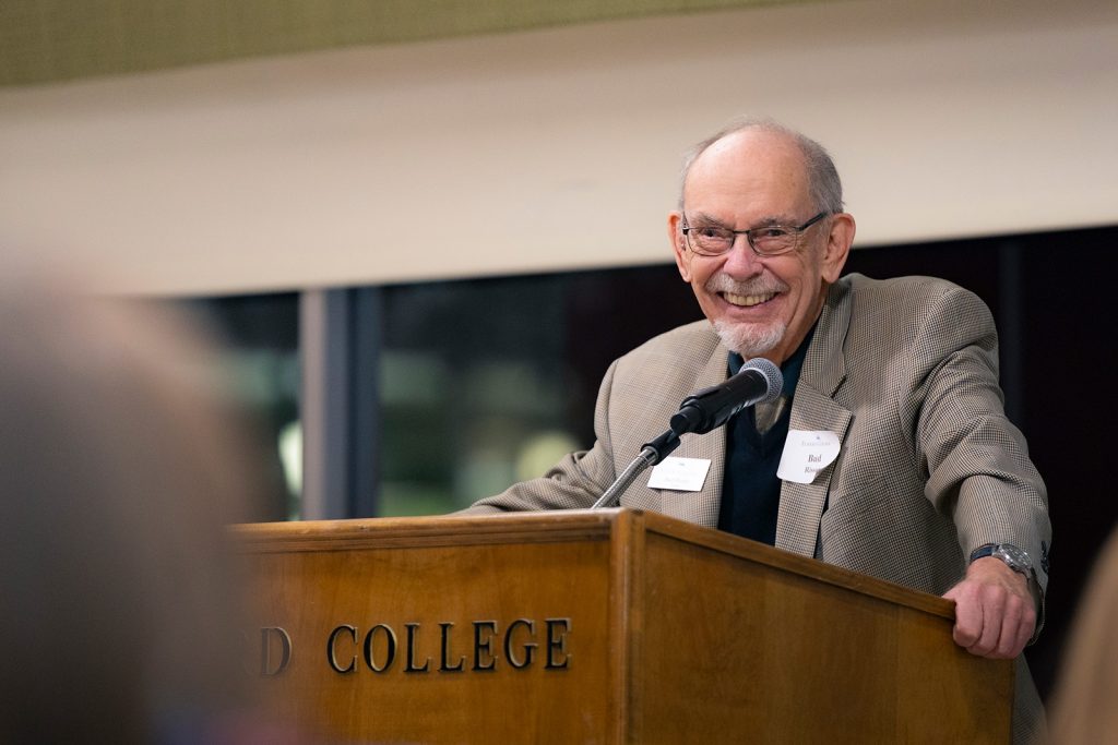 Gentleman at podium in suit and wearing glasses gives a big smile to the audience