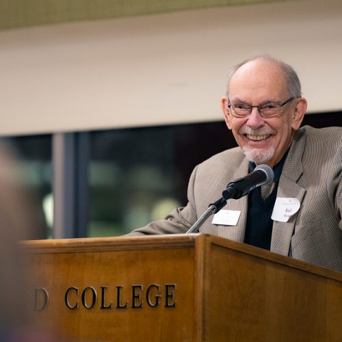 Gentleman at podium in suit and wearing glasses gives a big smile to the audience