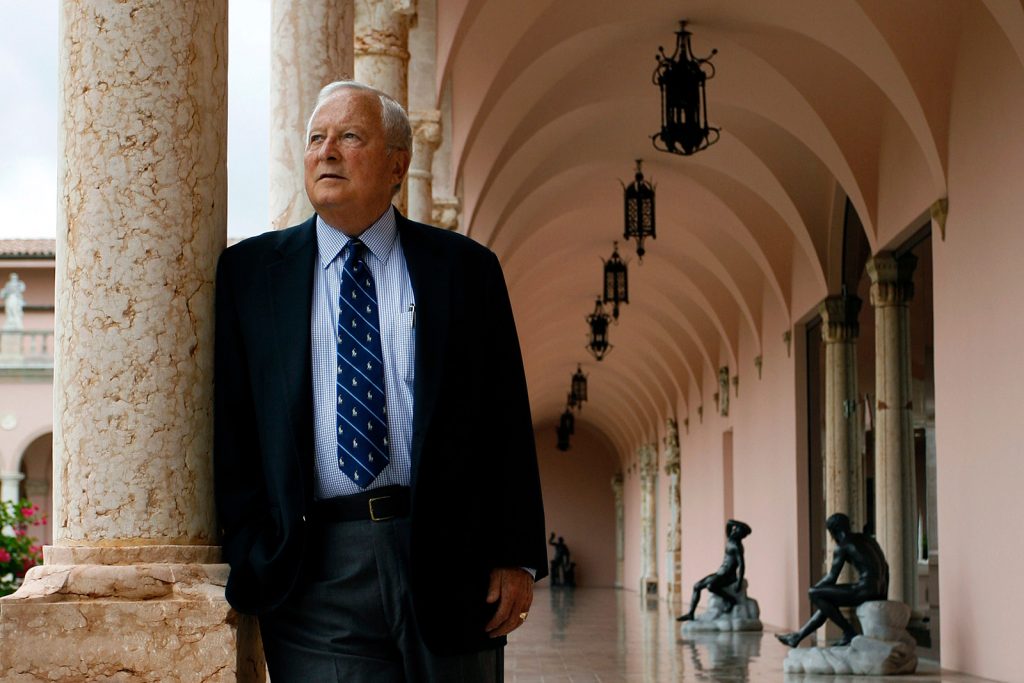 Man in suit and tie stands next to a marble column under an arcade lined with statues