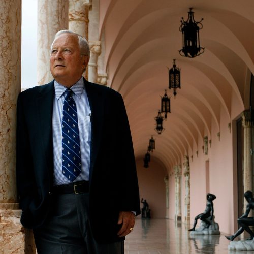 Man in suit and tie stands next to a marble column under an arcade lined with statues