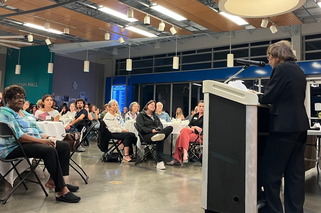 Woman standing at poidum speaks to a large group of people in a large room with hanging lights