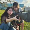 Two students squat behind a camera on a tripod with a very large lens as sun sets behind a pond behind them