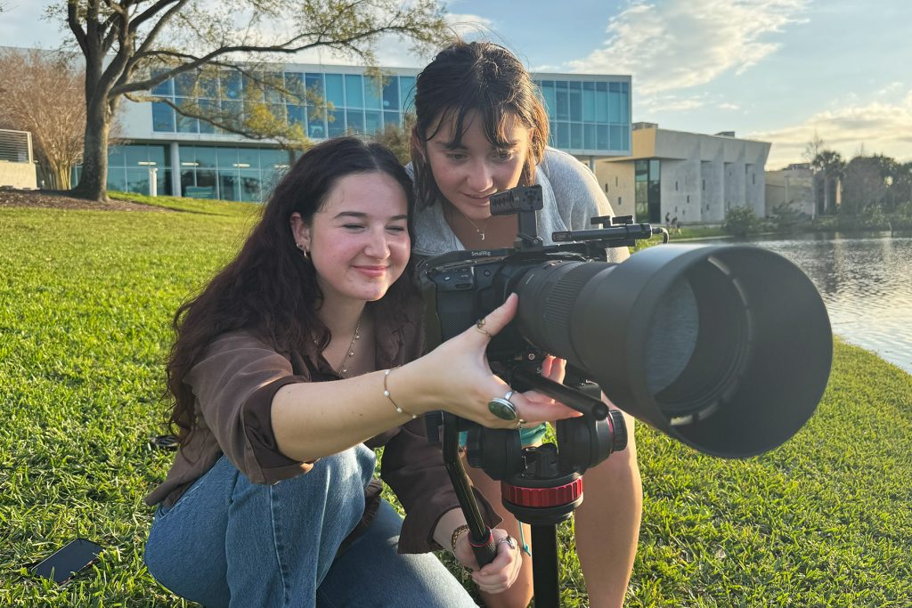 Two students squat behind a camera on a tripod with a very large lens as sun sets behind a pond behind them
