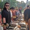 Students line both sides of a table covered with food at an outdoor setting
