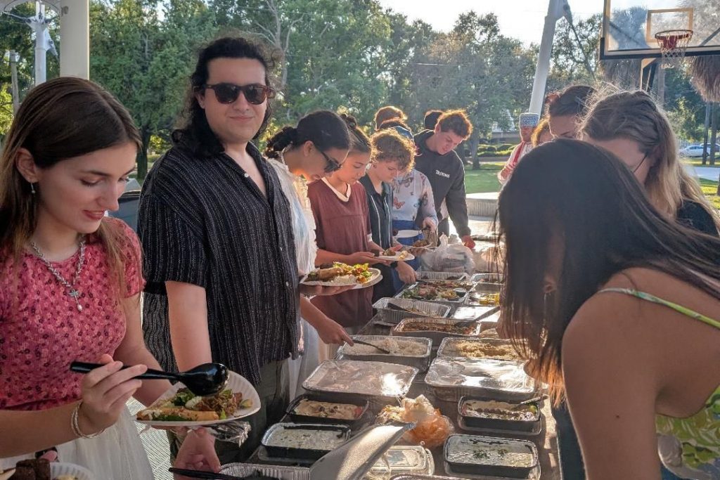 Students line both sides of a table covered with food at an outdoor setting
