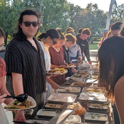 Students line both sides of a table covered with food at an outdoor setting