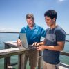 Professor and young man work on a laptop on a dock by the water.